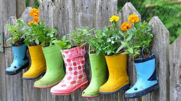 A variety of rainboots hung on a wooden picket fence with plants and salad greens planted in them.