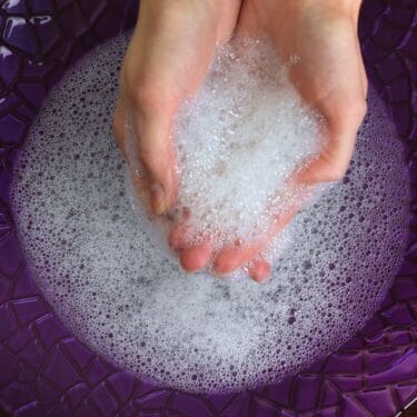 Hands scooping up dish detergent foam from a purple bowl.