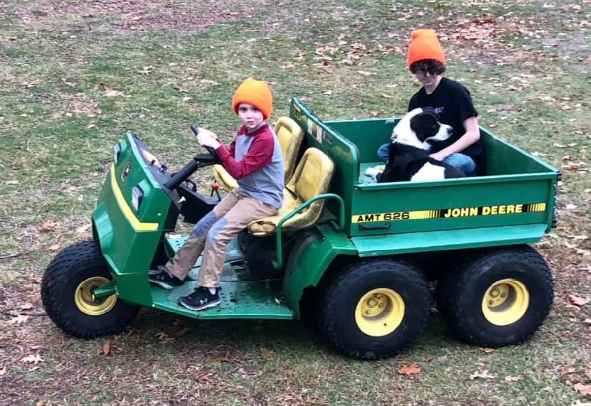 Two children wearing orange hats driving a John Deere AMT 626 with their black and white dog riding in the back.