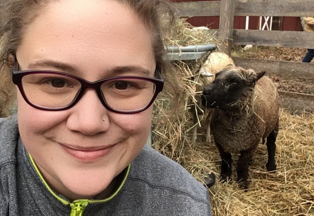 Jessica Knowles smiling in front of a sheep at her farm, surrounded by hay and wooden fencing.