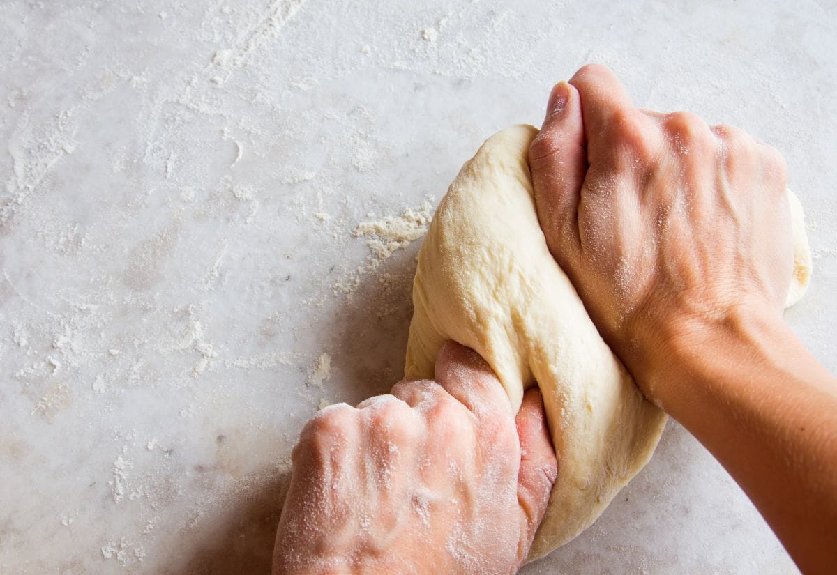 Hands kneading bread dough on a lightly floured countertop.