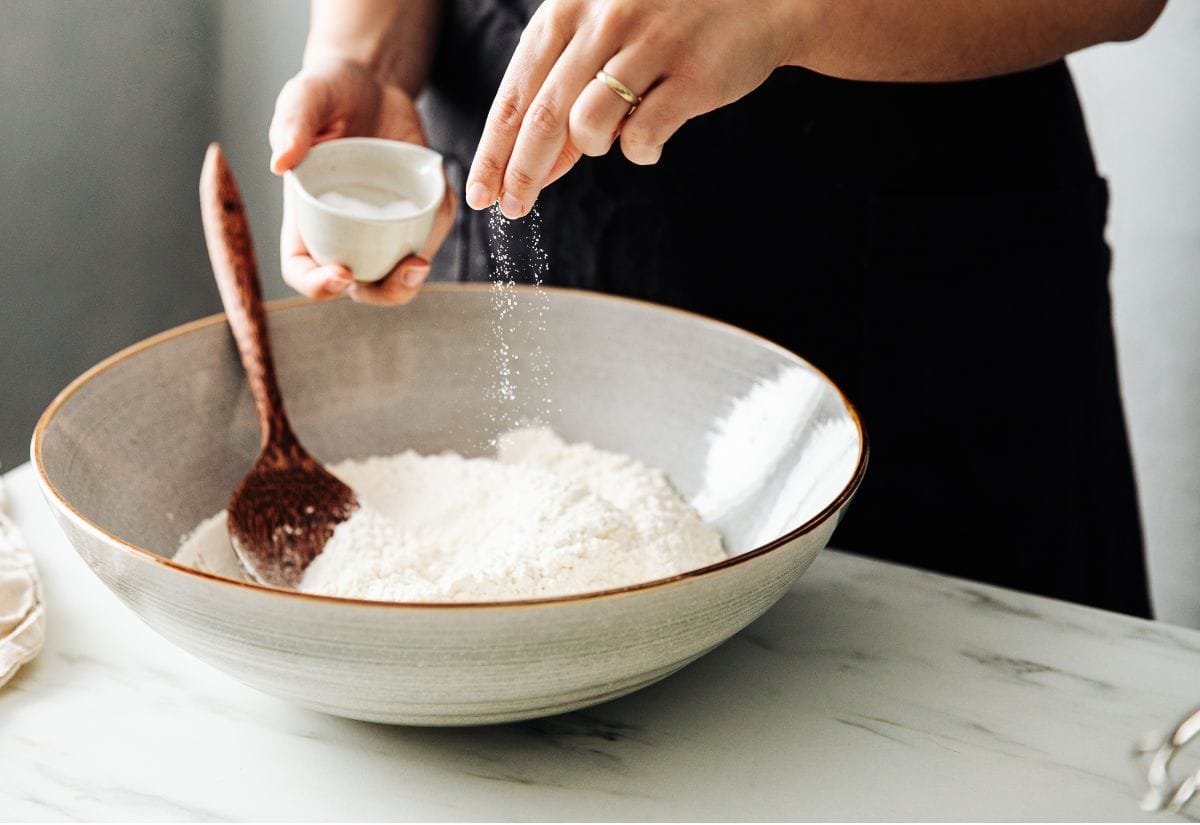 A person sprinkling salt into a bowl of flour with a wooden spoon.