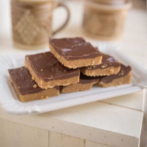 A stack of homemade peanut butter bars with a thick layer of milk chocolate on top, served on a white plate with mugs in the background.
