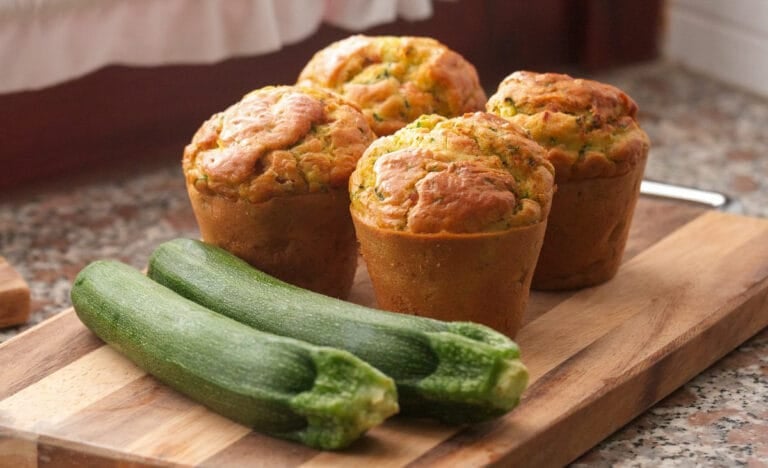 Four golden brown zucchini muffins on a wooden cutting board next to two whole zucchinis, set on a kitchen counter in warm light.