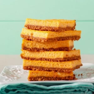 Stack of vibrant orange squash cheesecake bars with a graham cracker crust on a white plate with a turquoise napkin.