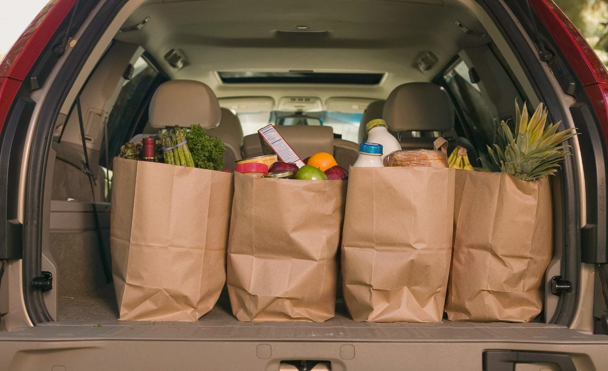 Paper grocery bags filled with fresh produce, bread, milk, and fruit lined up in the open trunk of a car after a grocery trip.