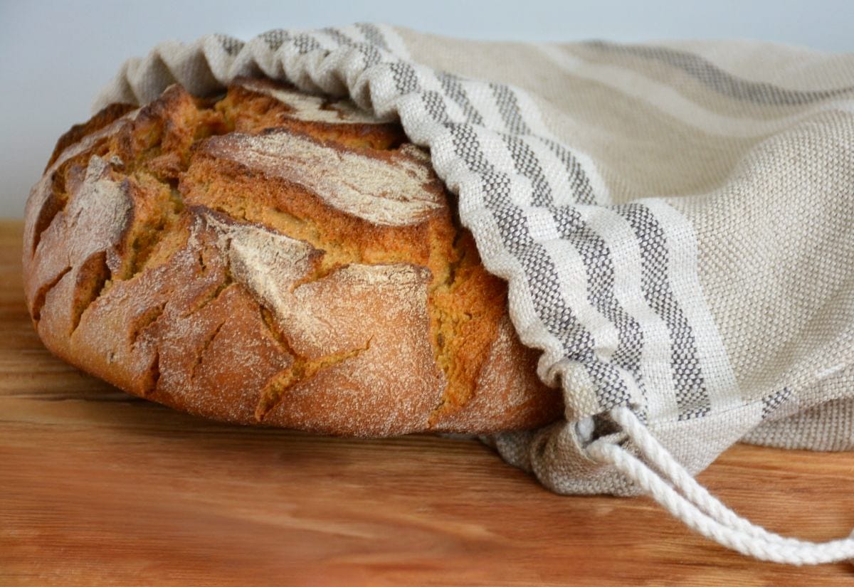 A rustic loaf of homemade bread partially wrapped in a linen bread bag on a wooden cutting board for breathable storage.