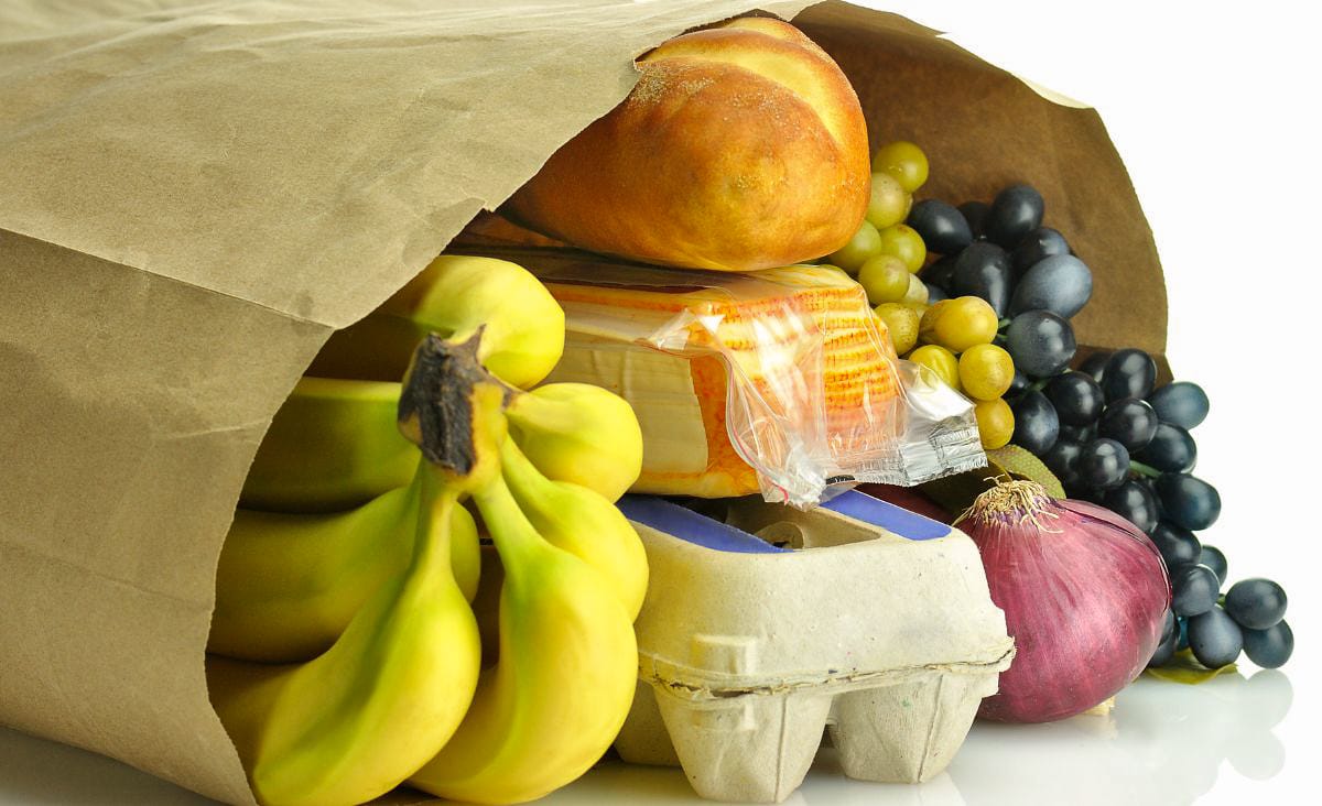 Brown paper grocery bag filled with fresh produce and staples including bananas, grapes, a red onion, a loaf of bread, an egg carton, and a block of cheese.