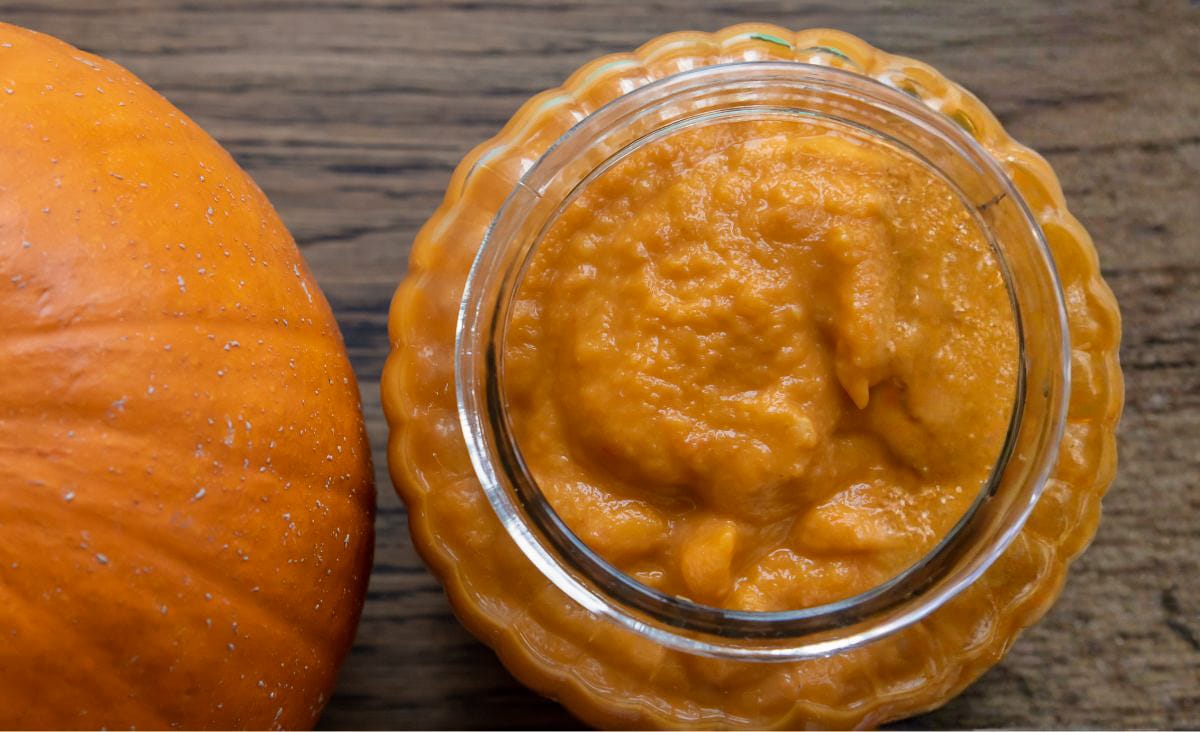 Top-down view of homemade pumpkin puree in a scalloped glass dish beside a whole pumpkin, showing the smooth, rich texture people can expect from fresh pie pumpkins.