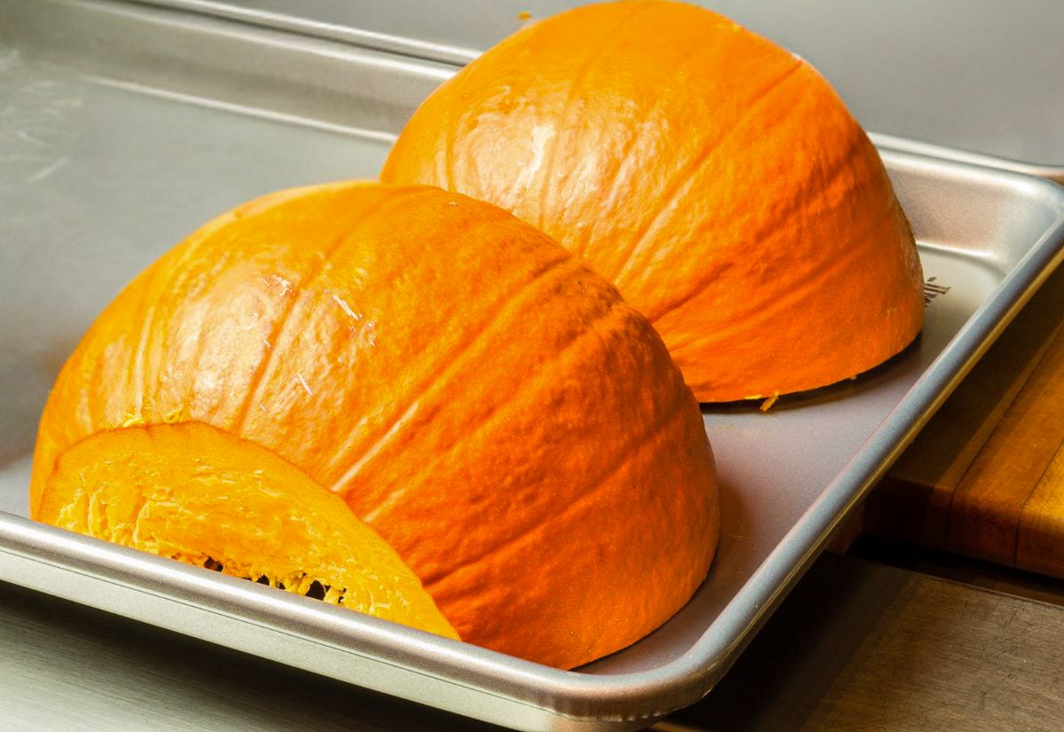 Two roasted pumpkin halves sitting cut-side down on a metal baking tray, showing the deep orange color and tender texture after oven roasting.