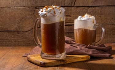 Two clear glass mugs of hot butterbeer, topped with whipped cream and dripping caramel, set on a wooden table with a brown napkin.