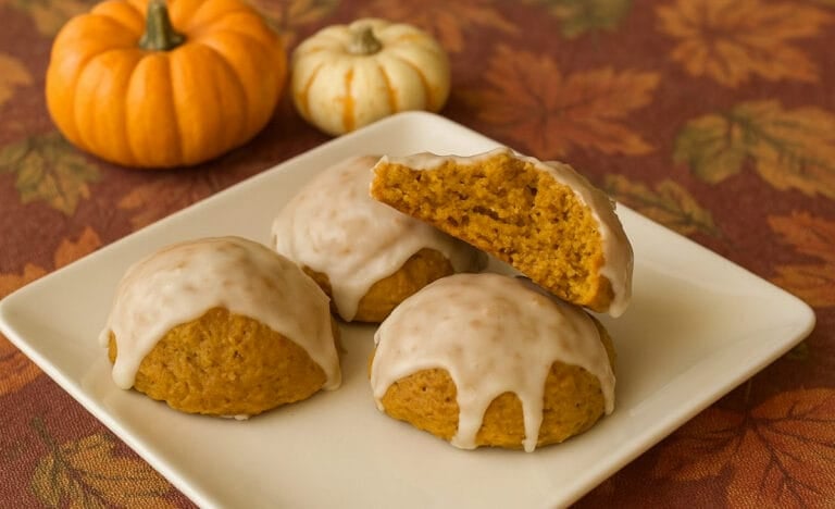 Four pumpkin cookies topped with maple glaze sit on a white plate with small ornamental pumpkins in the background. The cookies look soft and thick, with one broken open to reveal their chewy, golden-orange center against a fall-themed tablecloth.