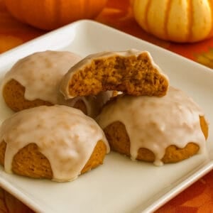 Close-up of three pumpkin cookies covered in maple glaze on a white plate, with one broken in half to show the soft interior. Small pumpkins and a fall-colored tablecloth in the background create a warm, autumn feel.