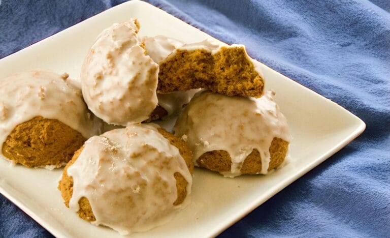 A plate of chewy pumpkin cookies drizzled with a glossy maple icing, with one cookie broken in half to show the soft interior.