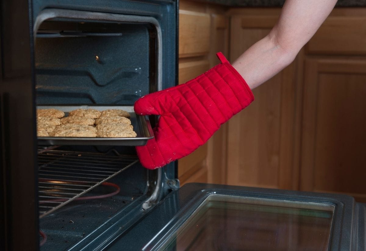 A person wearing a red oven mitt pulling a tray of freshly baked cookies out of the oven.