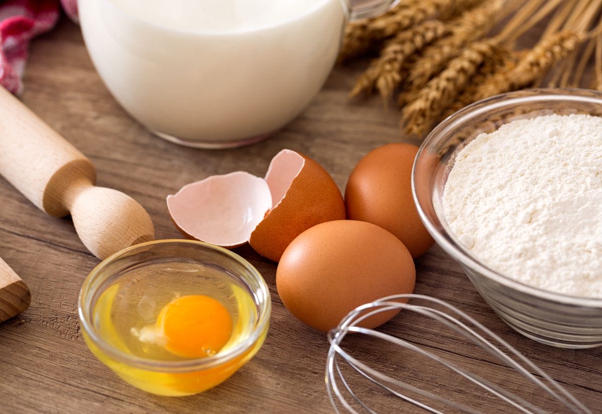 Fresh brown eggs, flour, milk, and baking tools on a wooden surface.