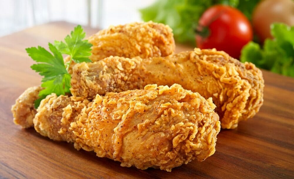 Fried chicken on a wooden cutting board with fresh veggies in the background.