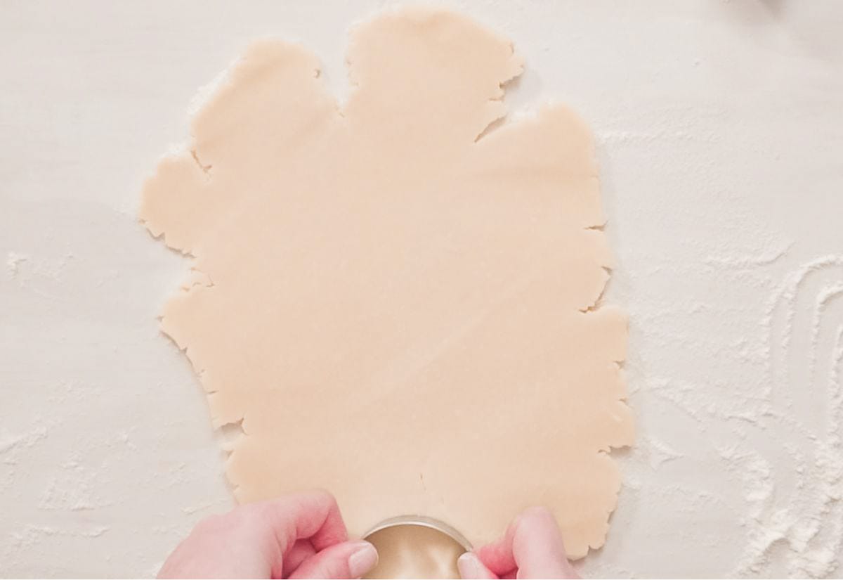 Hands pressing a round cookie cutter into rolled sugar cookie dough on a lightly floured surface.