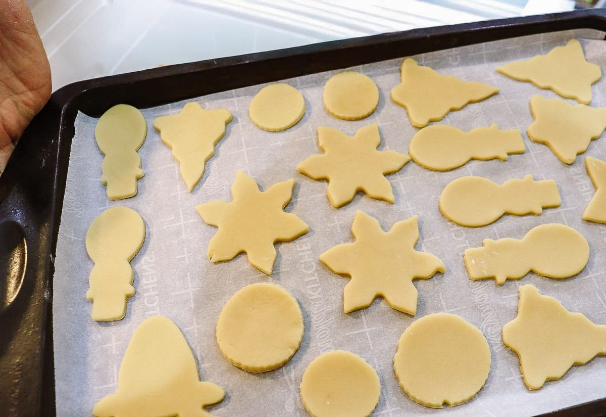 Unbaked cut-out sugar cookies in holiday shapes—snowflakes, bulbs, circles, and trees—arranged neatly on a parchment-lined baking sheet.