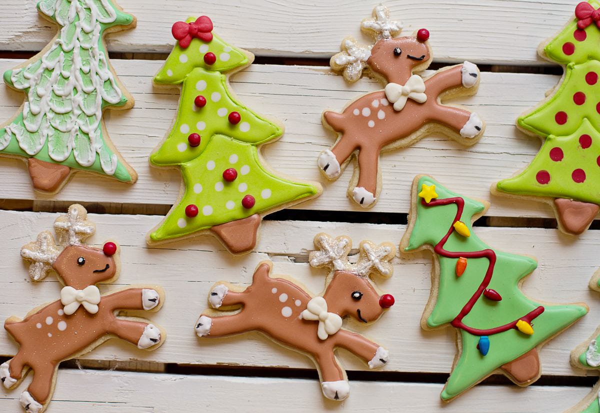 Decorated holiday sugar cookies shaped like reindeer and Christmas trees, frosted with bright natural icing in greens, reds, and browns.