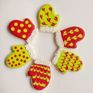 A ring of mitten-shaped sugar cookies decorated in red and neon green with polka dots, heart swirls, and feathered icing designs made with natural colors.