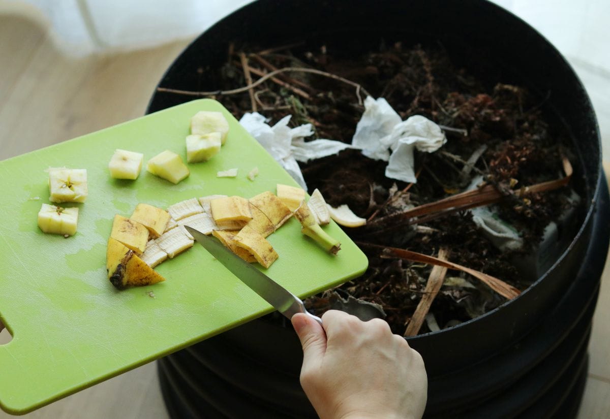 A person chopping a banana on a green cutting board over a compost bin filled with organic scraps.