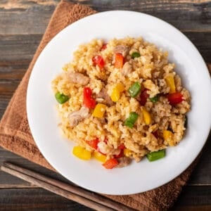 A white plate filled with colorful fried rice made with chopped red, yellow, and green bell peppers, rice, and pieces of pork, set on a brown towel with chopsticks nearby on a rustic wooden table.