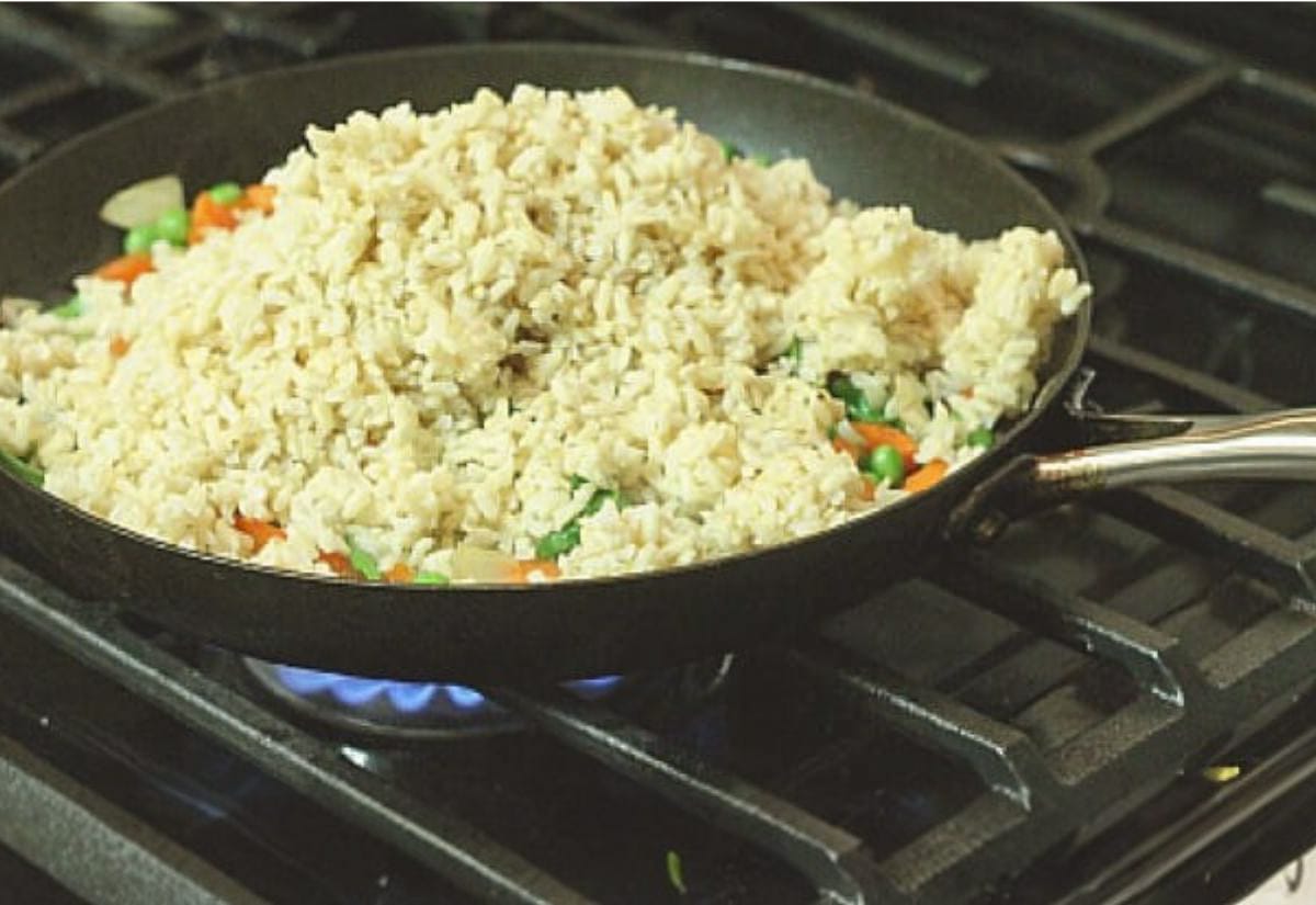 Skillet of fried rice being cooked on a gas stovetop, showing a mound of cooked brown rice on top of vegetables including peas, carrots, and onions.