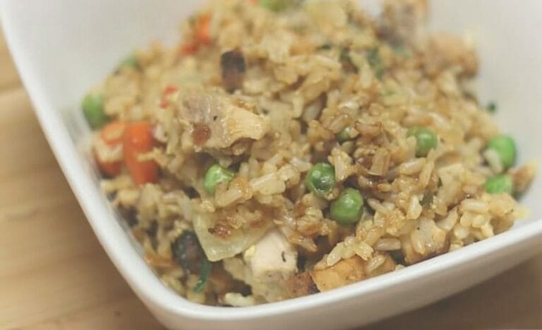 A white bowl filled with fried rice featuring visible chunks of pork, green peas, diced carrots, and caramelized onions.