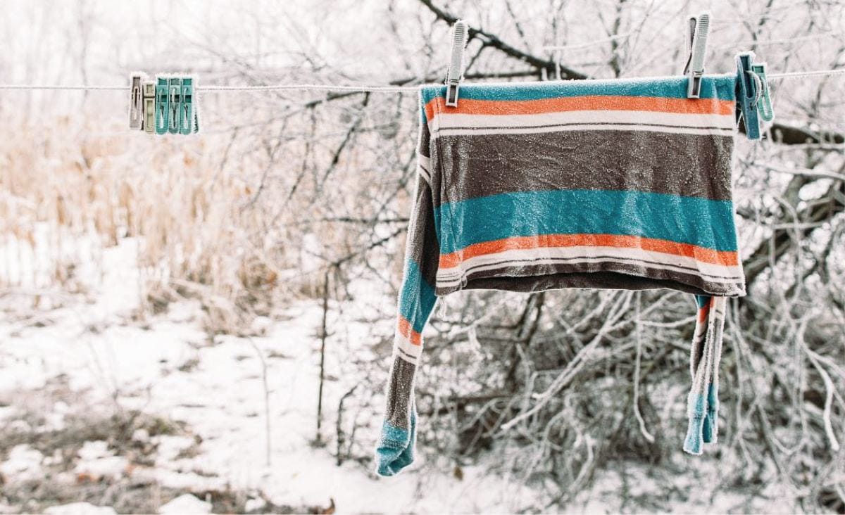 A striped top clipped to a clothesline in a snowy backyard, lightly frozen from the cold, with frosted branches and winter brush behind it.
