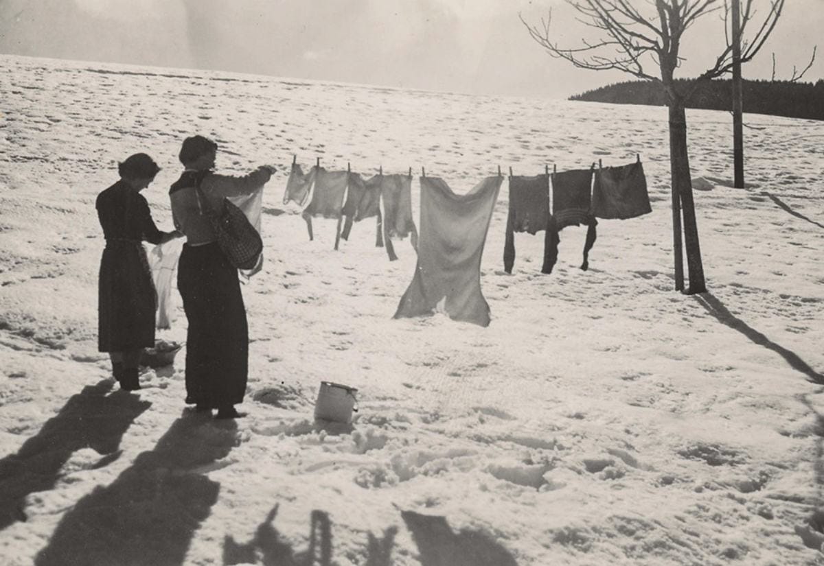 A vintage black-and-white photo of two women hanging laundry on a clothesline in a snowy field, surrounded by snow-covered ground and bare trees.