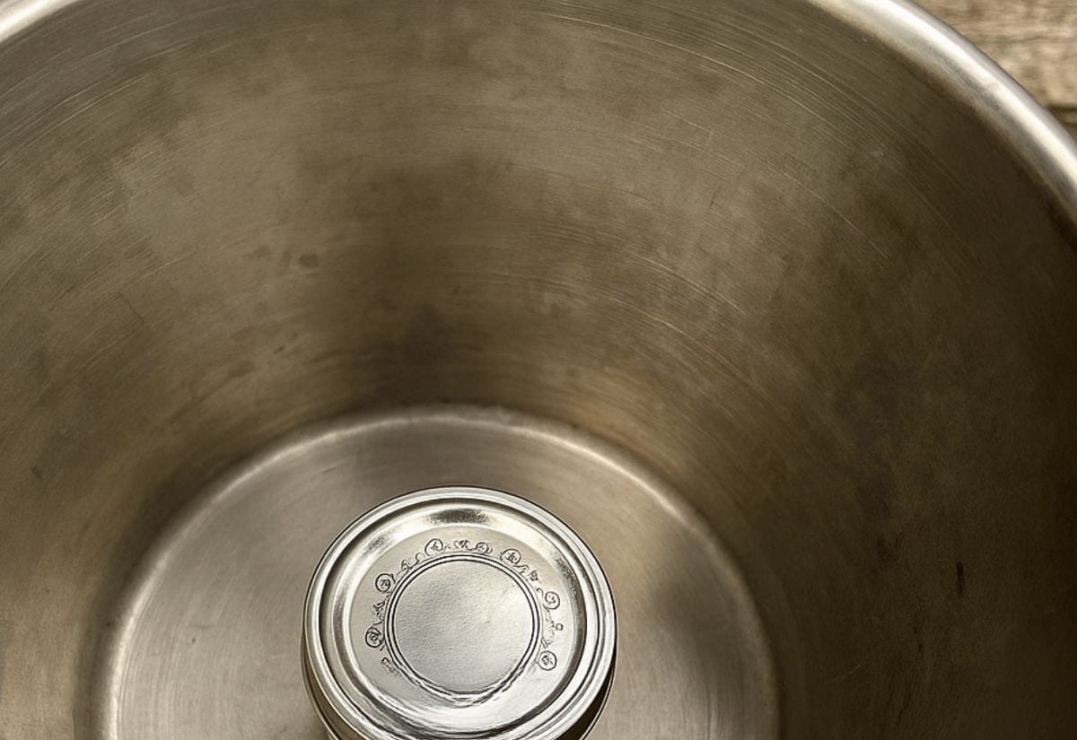 Close-up of a small frozen jelly jar sitting in the bottom of a stainless steel milking pail. The photo was taken in natural farmhouse light, showing the simple setup used to chill goat milk as it’s collected.