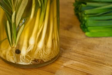 Close-up of a glass jar filled with wild onions and sprigs of rosemary submerged in golden pickling brine on a wooden countertop, with chopped greens in the background.