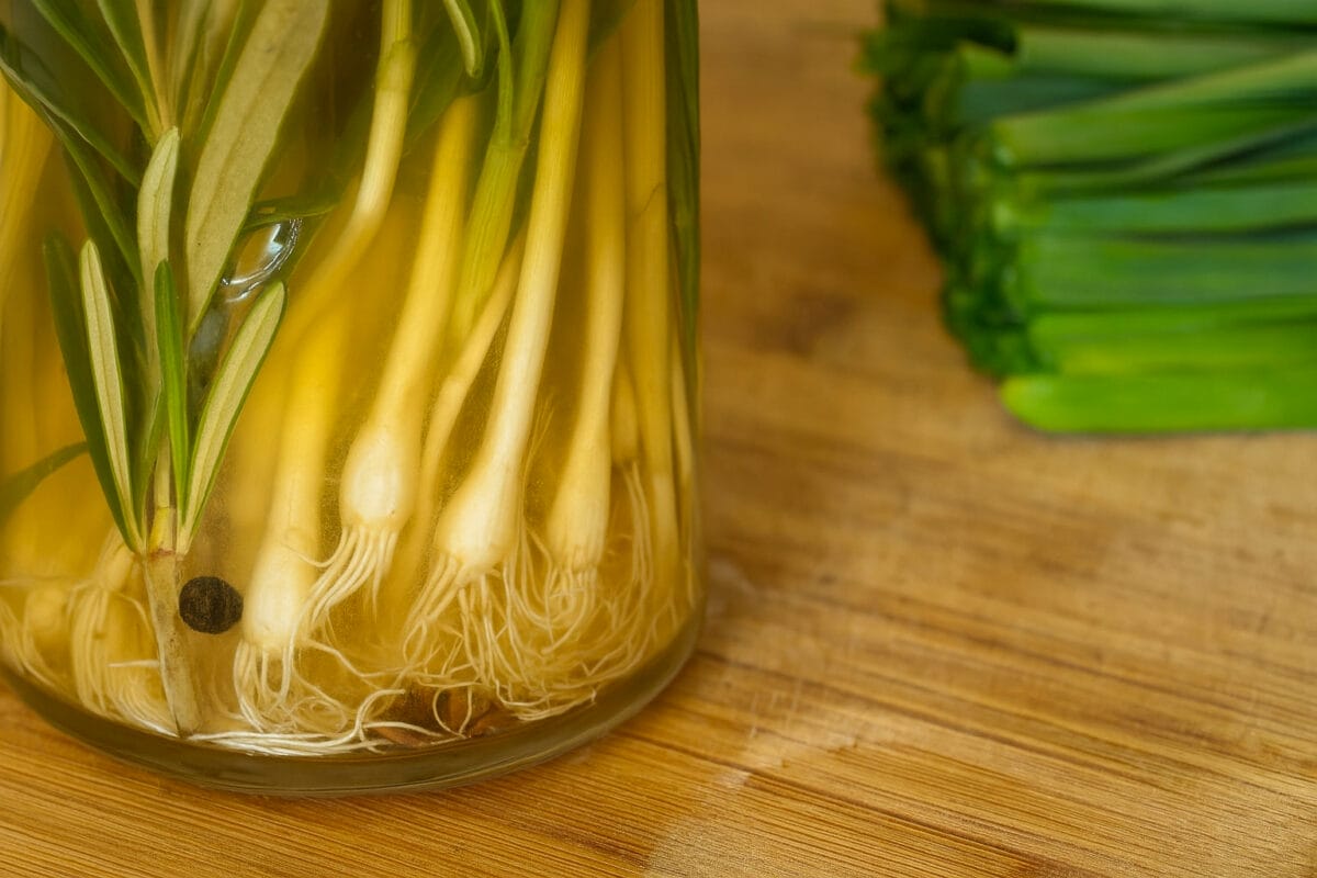 Close-up of a glass jar filled with wild onions and sprigs of rosemary submerged in golden pickling brine on a wooden countertop, with chopped greens in the background.