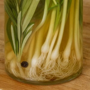 Square image showing a detailed view of wild onions with roots and stems submerged in brine alongside rosemary and peppercorns, inside a glass jar on a butcher block.