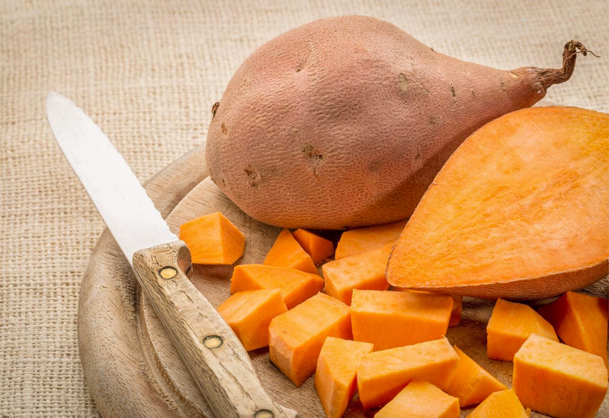 Fresh sweet potatoes on a wooden cutting board beside a knife, one whole and one cut into cubes, ready for prepping a homestyle stuffing recipe.