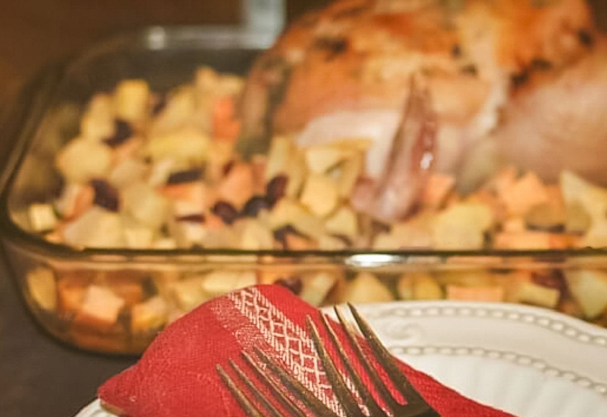 A serving scene with a white plate, red napkin, and fork in the foreground, and a baking dish of roasted chicken and sweet potato-apple stuffing blurred in the background — warm, inviting table setting.