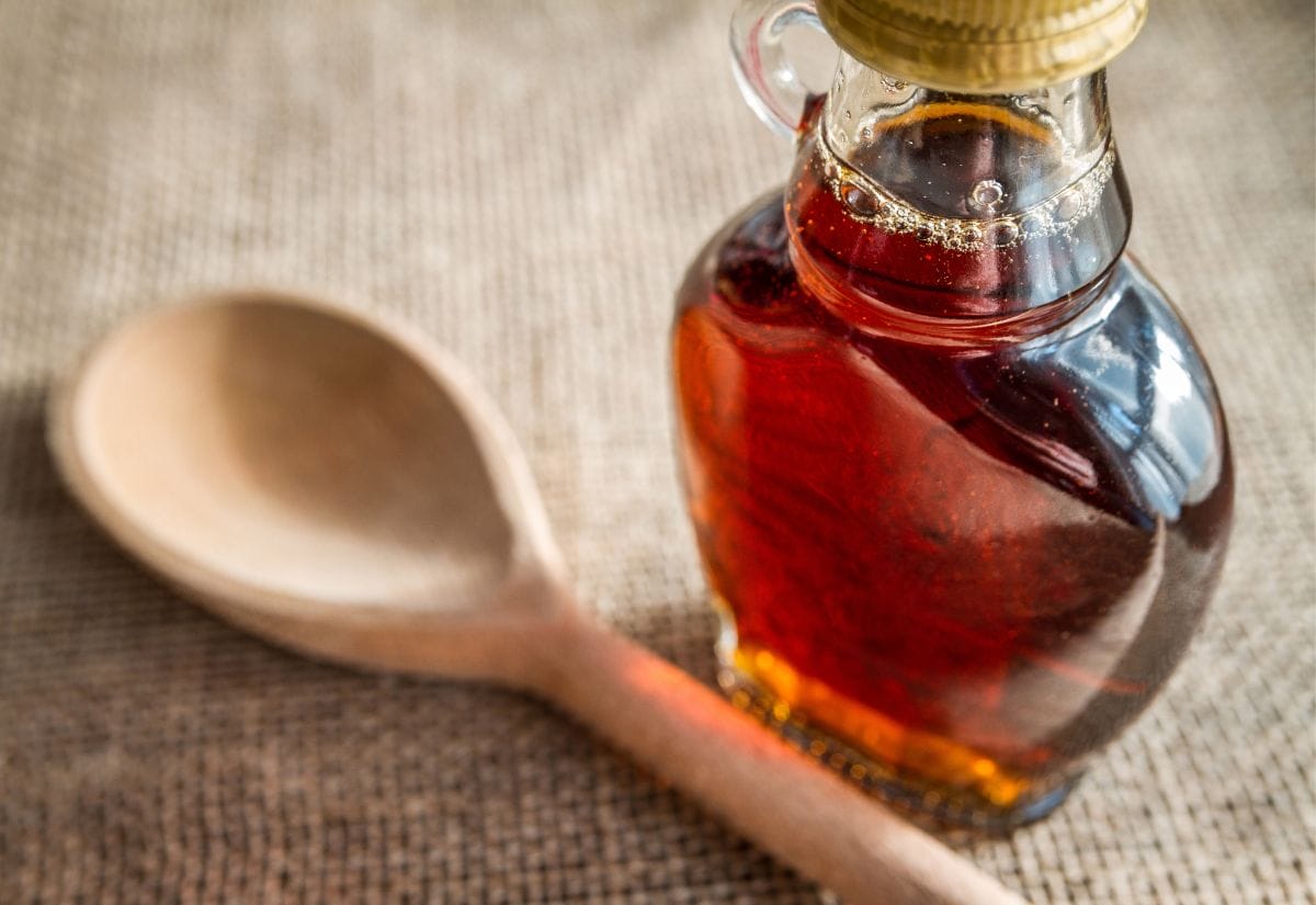 Glass bottle filled with golden syrup beside a wooden spoon on a burlap surface.