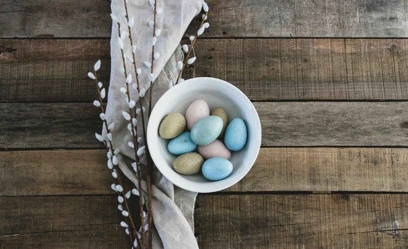 A white bowl on a wooden table filled with naturally dyed easter eggs.