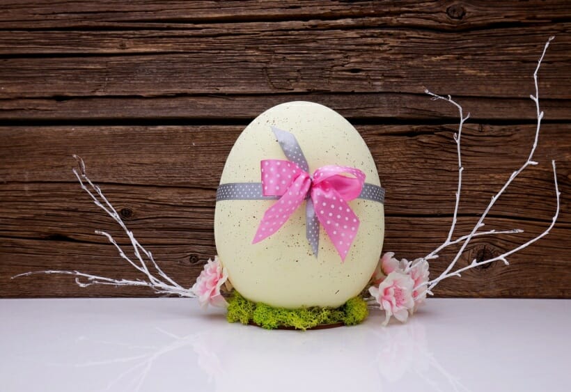 A pale yellow easter egg sitting on moss with a pink polka dot bow.