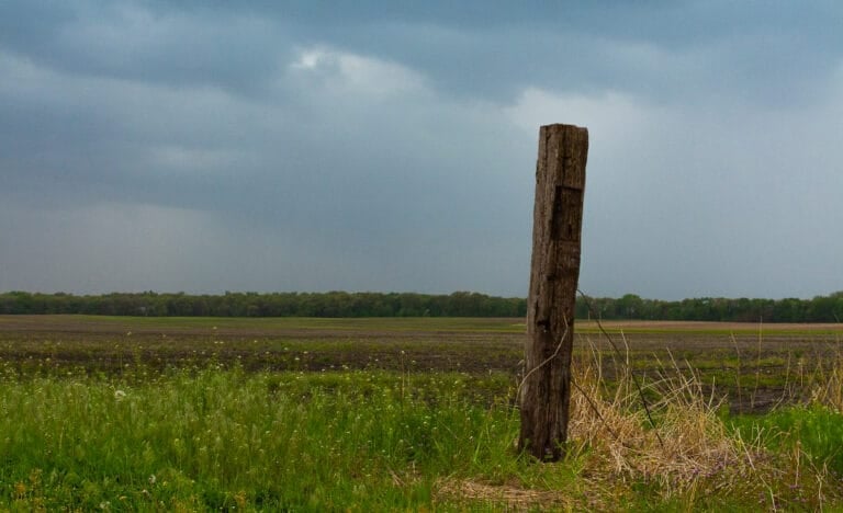 A wide view of an open farm field beneath a dark, cloudy sky, with a single weathered wooden fence post in the foreground and trees lining the horizon.