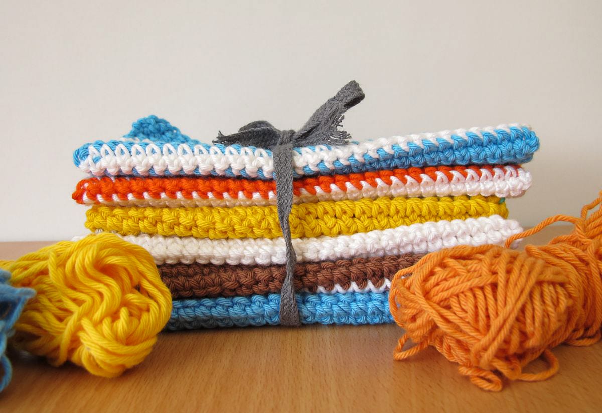 A bundle of neatly stacked, handmade crocheted dishcloths in bright colors tied with a gray ribbon, surrounded by balls of yarn on a wooden surface.
