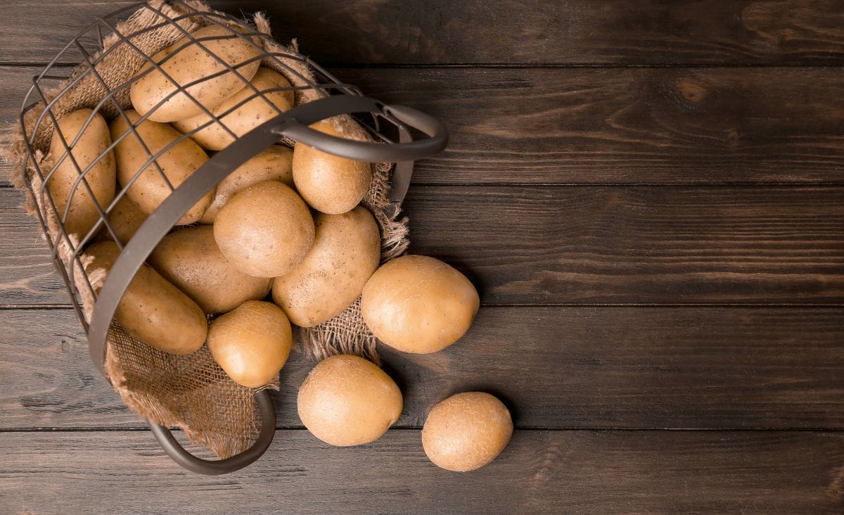 A wire basket of potatoes spilling out on a wooden table.