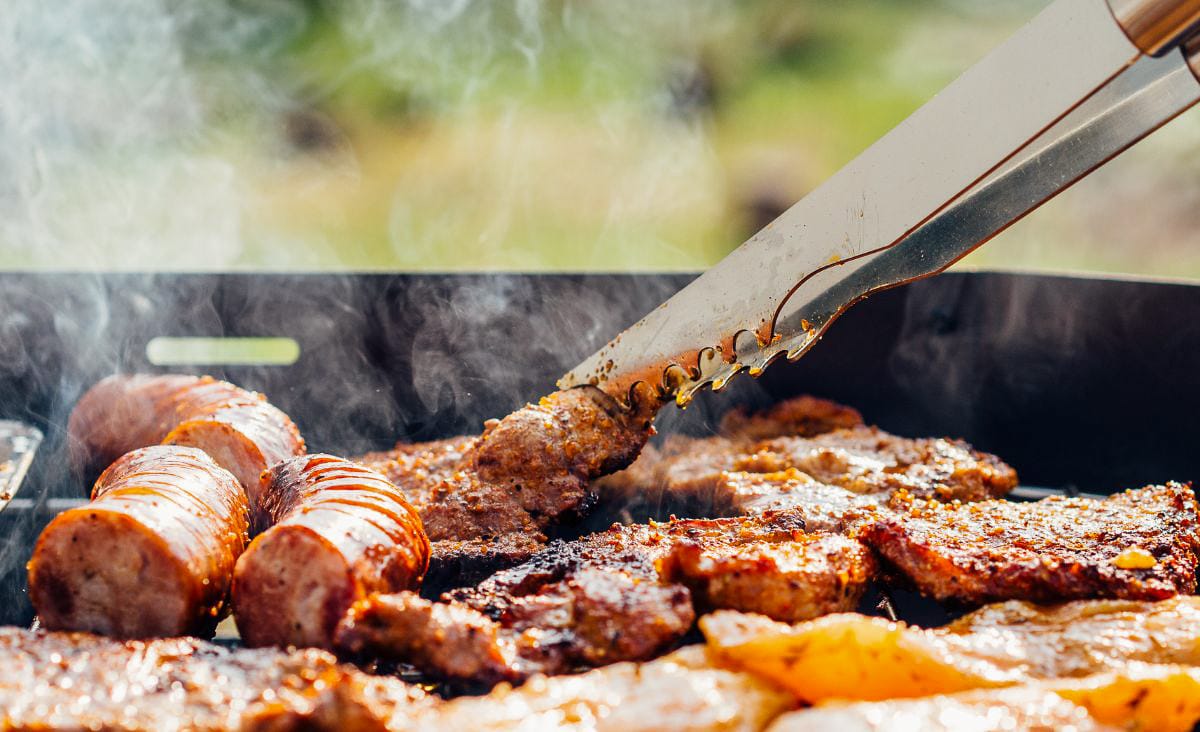 A variety of meats sizzling on a grill, with smoke rising and a pair of tongs flipping a steak.
