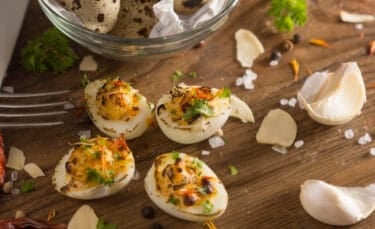 Overhead photo of herb- and pepper-topped deviled quail eggs on a rustic wooden surface with garlic cloves, peppercorns, and coarse salt scattered around.