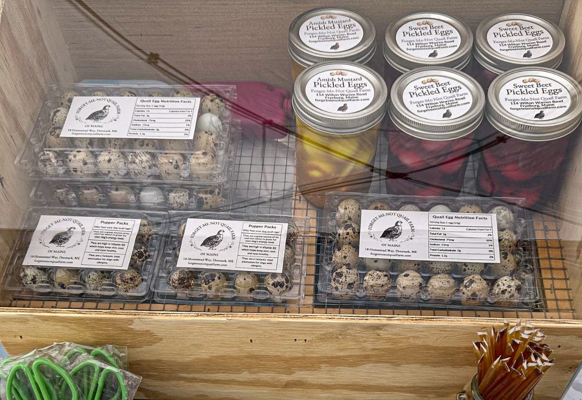 Market display showing cartons of fresh quail eggs and jars of pickled quail eggs labeled with flavors like Amish Mustard and Sweet Beet from Forget-Me-Not Quail Farm.