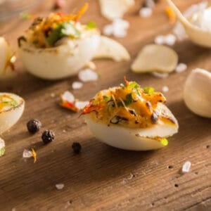 Square-format close-up of a single spicy deviled quail egg garnished with parsley and chili flakes, set on a wooden background with visible grains of sea salt and black pepper.