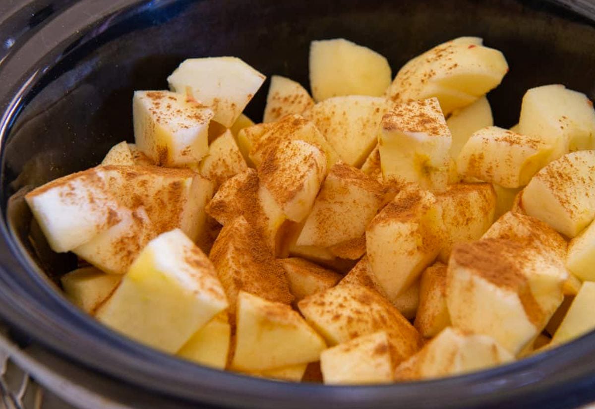 Peeled and chopped apples sprinkled with cinnamon inside a black crockpot, ready for slow cooking.