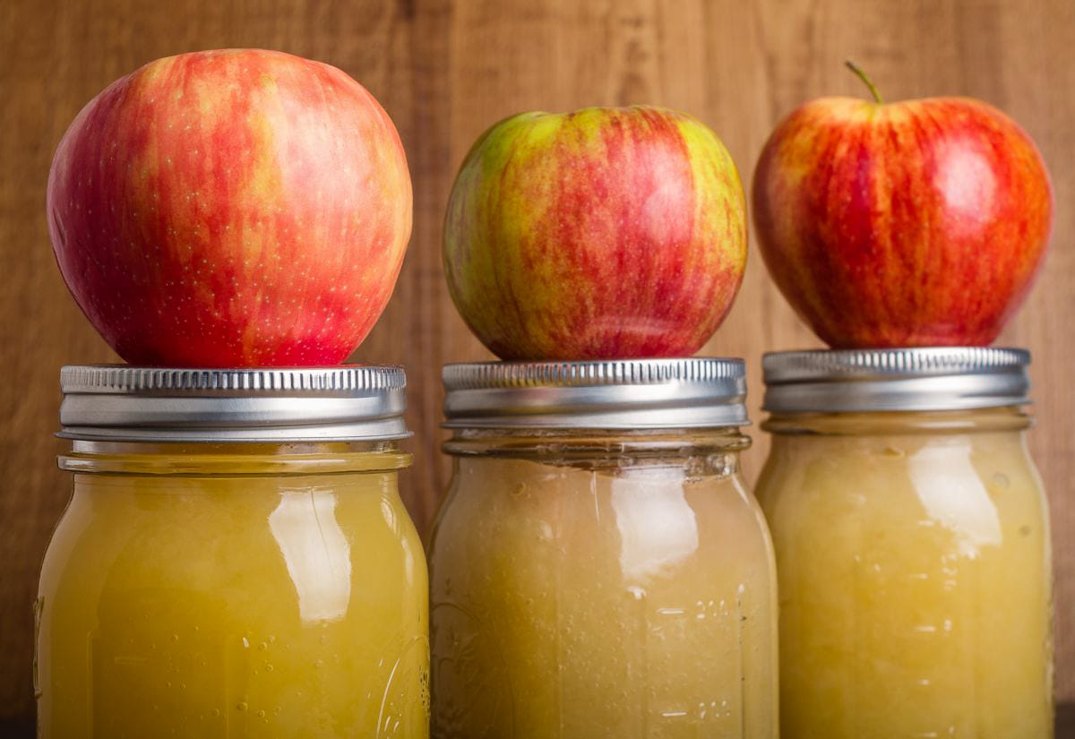 Three jars of canned homemade applesauce with red apples resting on top of the lids.