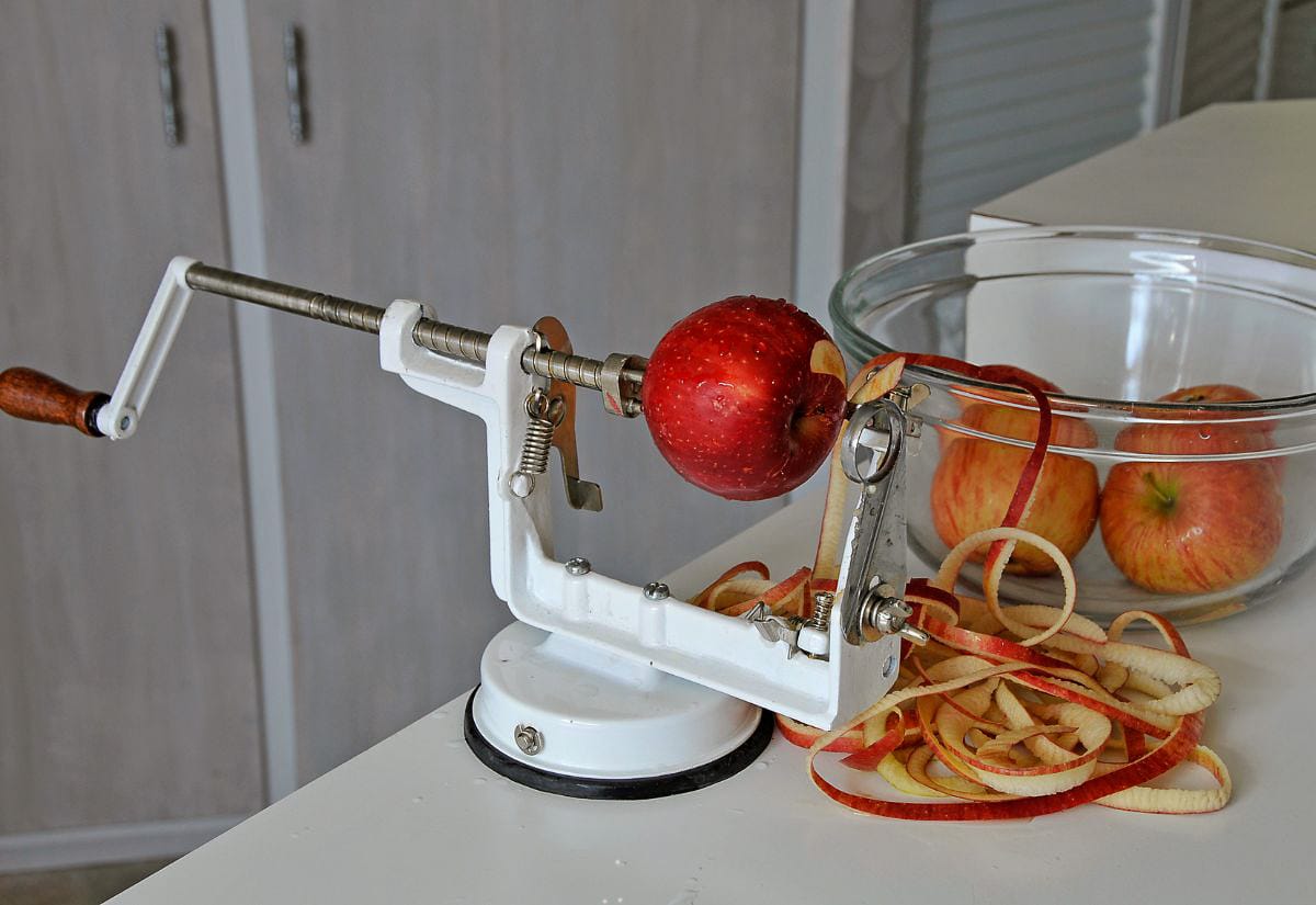 Hand-cranked apple peeler peeling an apple over a bowl filled with apples and apple peels.